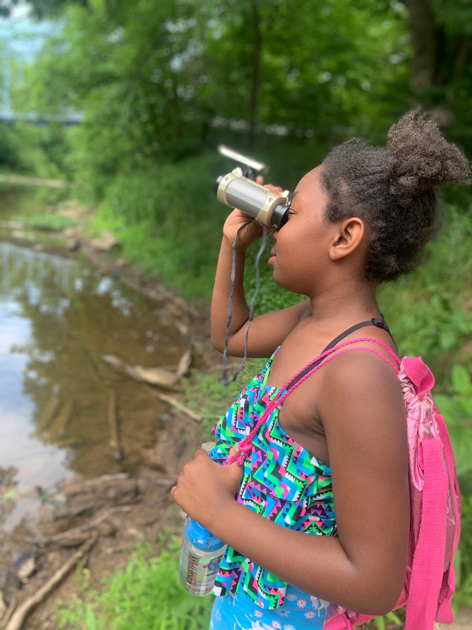 Young girl wearing a pink drawstring bag and holding binoculars by the edge of the water at Camp Cann-Edi-On